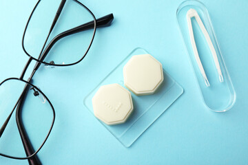 Contact lens case, glasses and tweezers on light turquoise background, flat lay