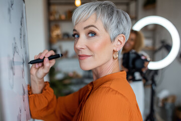 A creative professional with short silver hair engages in a brainstorming session, writing on a whiteboard in a bright studio filled with artistic decor and natural light