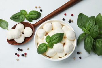 Tasty mozzarella cheese balls in bowl, spoon, basil and peppercorns on white marble table, flat lay