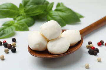 Tasty mozzarella cheese balls in spoon, basil and peppercorns on white marble table, closeup