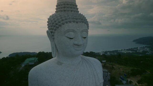 Phuket Big Buddha statue head against tranquil ocean under cloudy sky aerial view. Buddhist god radiates calmness and tranquility at sunrise. Spellbound architectural monument