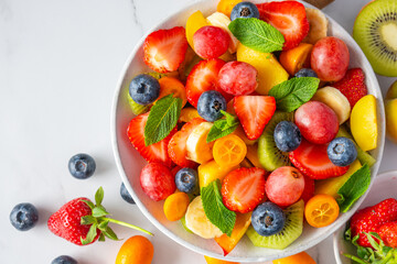 Summer colorful fruit salad in a bowl on white marble background. Top view with copy space. Healthy food for breakfast. Mixed strawberries, grapes, banana, kiwi, blueberries, peach, citrus