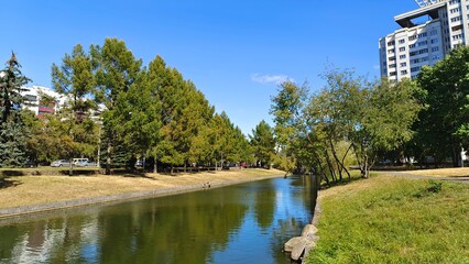 Spruce, larch and other trees grow on the grassy concrete banks of the canal. There are buildings, a roadway and walking paths nearby. Cars drive by. Ducks swim in the water. Sunny autumn weather