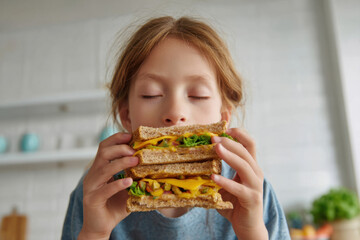 Young girl is happily eating an open-faced sandwich topped with fresh lettuce and melted cheese. She appears content and focused on her meal in a cozy kitchen