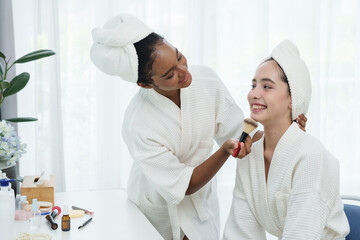 African American teenage girl gently applying makeup on smiling Caucasian Asian teenage friend in spa robes, expressing beauty routine, confidence, connection and multicultural friendship