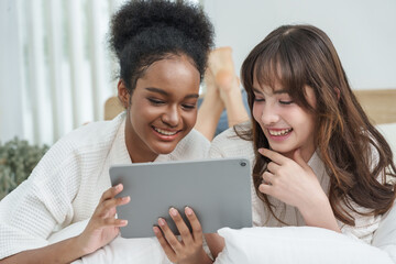 Two teenage girls lying on bed smiling at tablet screen, African American and mixed-race Caucasian Asian friends sharing laughter, relaxed digital lifestyle and joyful bonding time together