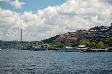 Fishing boats on the Bosphorus with large ships passing under the Bosphorus Bridge in Istanbul, Turkey on a bright day with scenic clouds, hills, and a Turkish flag in the background