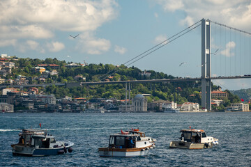 Fishing boats on the Bosphorus with large ships passing under the Bosphorus Bridge in Istanbul, Turkey on a bright day with scenic clouds, hills, and a Turkish flag in the background.