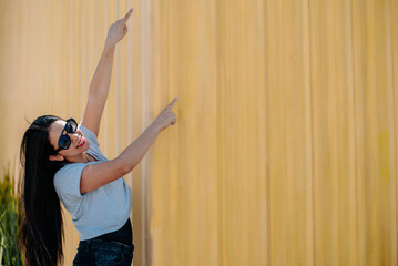 Joyful young woman with sunglasses pointing up, standing against a yellow wall in a dynamic pose.