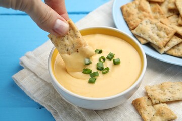 Woman dipping cracker into tasty cheese sauce at light blue wooden table, closeup