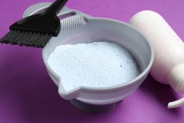 Bowl with hair bleaching powder, bottle of developer and brush on purple background, closeup