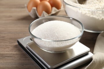 Digital kitchen scale with bowl of sugar and other products on wooden table, closeup