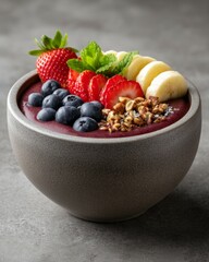 A smoothie bowl topped with fresh strawberries, blueberries, banana slices, granola, and mint leaves, presented in a matte gray bowl on a textured gray surface.