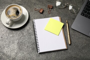 Notebook, pen, sticky note, coffee, binder clips, glasses and laptop on grey table, above view