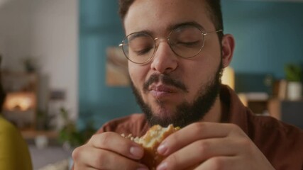 Close-up shot of young smiling man eating small burger and sipping drink from plastic cup at home
