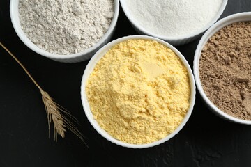 Different types of flour in bowls and spike on black table, flat lay
