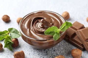 Tasty chocolate spread with mint in bowl and hazelnuts on grey textured table, closeup