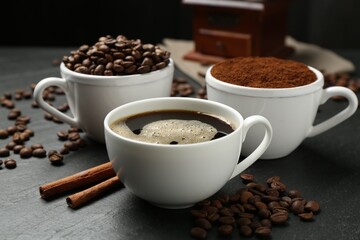 Cup of drink, ground coffee, beans and cinnamon on black table, closeup