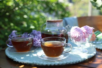 Aromatic tea in cups, teapot, zephyrs and lilac flowers on wooden table in garden, closeup