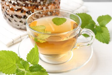 Aromatic lemon balm tea in glass cup, teapot and fresh leaves on white table, closeup