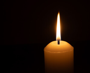 A single burning candle flame or light glowing on a big white candle isolated on black or dark background on table in church for Christmas, funeral or memorial service