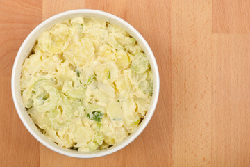 A white bowl filled with creamy potato salad mixed with scallions and celery sits on a light wooden surface