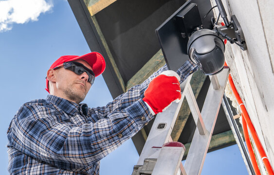 Technician Installs Exterior Security Camera on a Residential Building During Bright Day in a Suburban Neighborhood