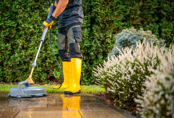 Fototapeta na wymiar Person Uses a Pressure Washer to Clean Patio Stones While Wearing Yellow Boots and Standing on a Well-Maintained Lawn Near Blooming Bushes During a Sunny Afternoon
