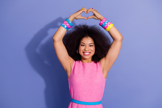 Joyful young woman in vibrant pink outfit forming heart symbol with hands, expressing happiness against a purple backdrop