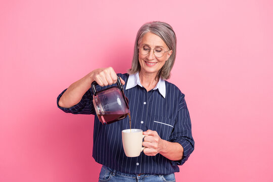 Content senior woman enjoying leisure time pouring fresh coffee into a mug with a cheerful smile on a pink backdrop