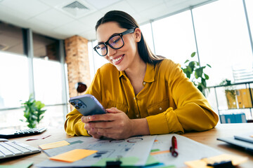 Young businesswoman browsing phone in modern office workspace with natural lighting and casual...