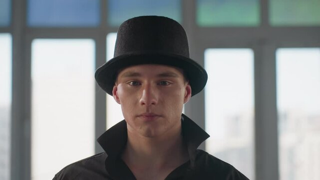 confident man in black shirt and hat tilting brim gazing at camera inside loft studio with large window and mirrored reflections showing neck tattoo under popped collar and subtle smile