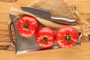 Three pink tomatoes with a metal tray and a knife on a wooden table, close-up, top view.
