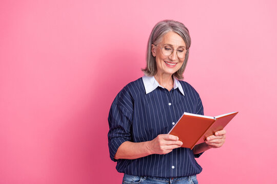 Confident older woman with glasses reads a book against a pink backdrop, showcasing charm and thoughtful expression