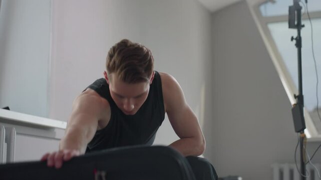 Young man in black tank top sits on stool by open window, adjusts black jacket on lap as sunlight filters through high rise building outside in minimalist modern apartment interior