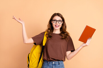 Young woman holding a book and gesturing happily with a yellow backpack in casual wear on beige...