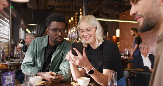 Multicultural people sitting at cafe. Multiethnic friends sitting at table. Cheerful group of people drinking coffee at restaurant. Smiling friends talking about photo at phone. Ethnic diversity.