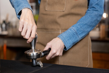 Close-up hands of Asian female barista pressing espresso grounds using tamper tool in coffee shop. Wearing brown apron and denim shirt, showing barista skill and professional brewing preparation.