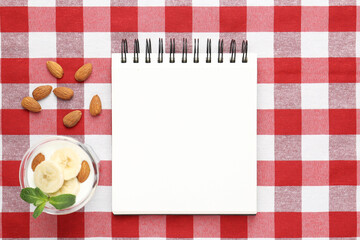 Blank recipe book, banana dessert and almonds on checkered tablecloth, top view. Mockup for design