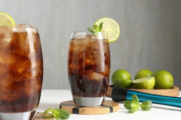 Tasty Cuba Libre cocktail in glasses, ingredients and muddler on white table