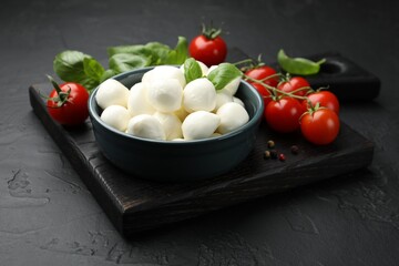 Tasty mozzarella cheese balls in bowl, basil, peppercorns and tomatoes on black table, closeup
