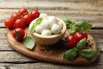 Tasty mozzarella cheese balls in bowl, basil, peppercorns and tomatoes on wooden table, closeup