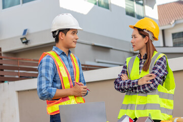 Male and female engineers wearing hard hats smile and discuss blueprints on a construction site, demonstrating teamwork, safety and expertise.
