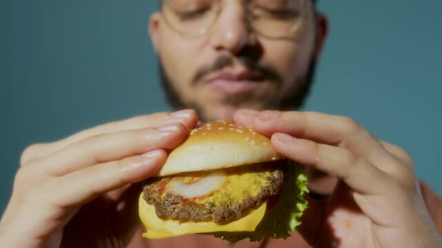 Close-up shot of young bearded man in glasses taking bite of delicious burger for fast food advertisement