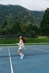 Obraz premium Beautiful Asian woman playing tennis on a sunny day, radiating joy and energy, surrounded by lush green nature and an inviting blue court