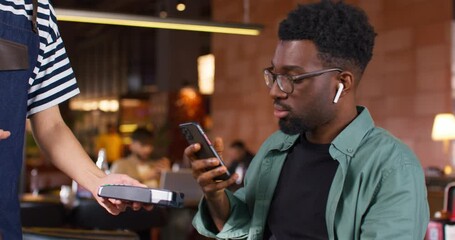 African American man conducting non contact payment. Man wearing glasses and wireless headphones. Smiling male spending work break at cafe. Handsome man in green shirt. Waiter and client.