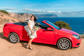 A woman in a white dress stands in front of a red convertible car. She is holding a bouquet of...