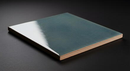 Close up of a single square teal tile with a glossy finish on a dark surface area