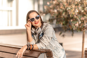 Fototapeta premium A young girl with long hair in jeans and headphones. Sitting on a bench.