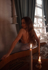 young brunette beautiful girl in blue slim dress stands sensual from the back near black grand piano with candles and looks apart on vintage room background with window. lifestyle concept, free space